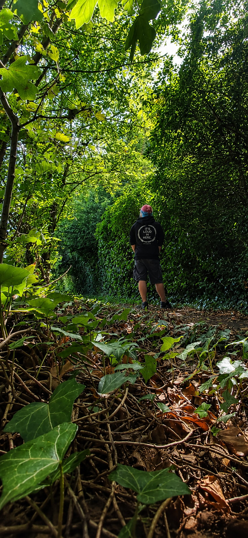 A women wearing a black hoodie with mental health awareness quote  on the back stands on a leaf-covered path in a dense green forest promoting mental health and neurodivergence awareness by muted mayhem