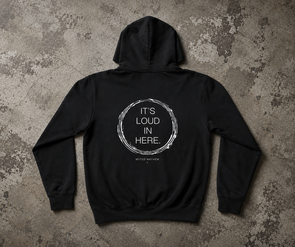 A black pullover hoodie laid flat on a textured gray concrete-style background, showing the back view with a white circular graphic and the text 'IT'S LOUD IN HERE.' printed on it. promoting mental health and neurodivergence awareness by muted mayhem