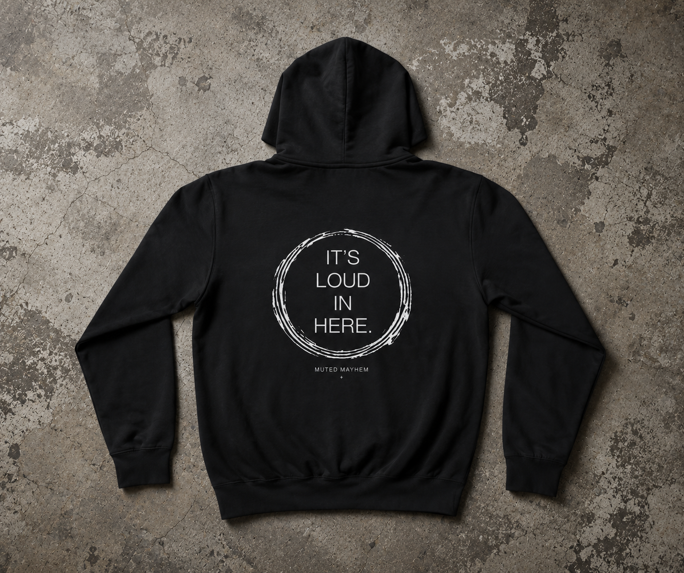 A black pullover hoodie laid flat on a textured gray concrete-style background, showing the back view with a white circular graphic and the text 'IT'S LOUD IN HERE.' printed on it. promoting mental health and neurodivergence awareness by muted mayhem