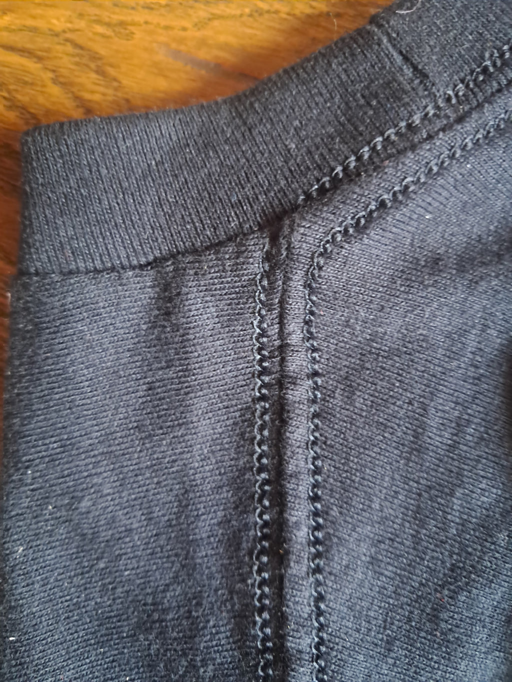 Close-up of a dark gray ribbed knit garment, showing the neckline and double-stitched seam details, with a wooden surface visible in the top-left corner promoting mental health and neurodivergence awareness by muted mayhem.