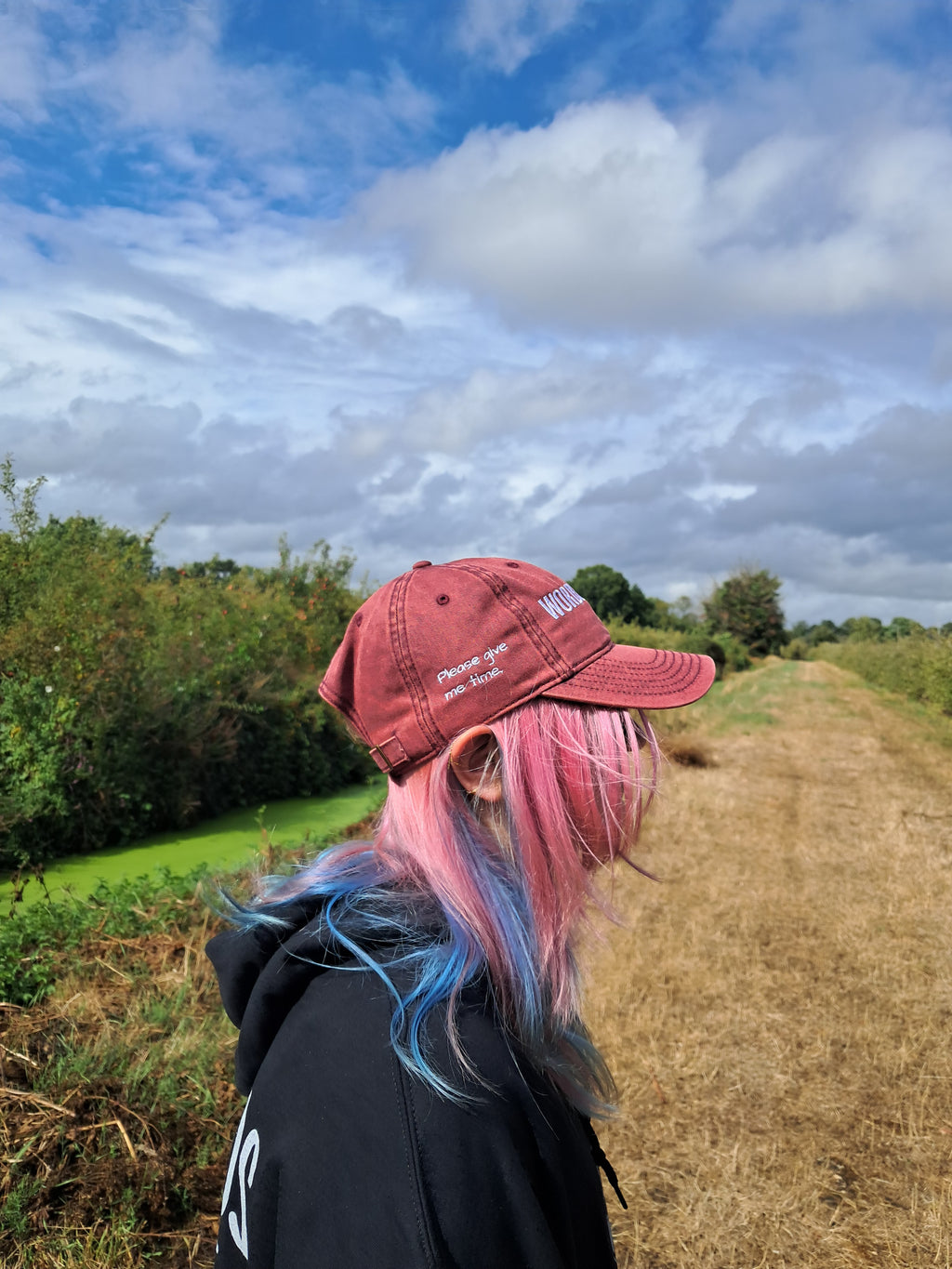 A person outdoors on a dirt path wearing a dusty red baseball cap with white embroidered text and pink-to-blue dyed hair, photographed from the side against a cloudy blue sky and greenery.