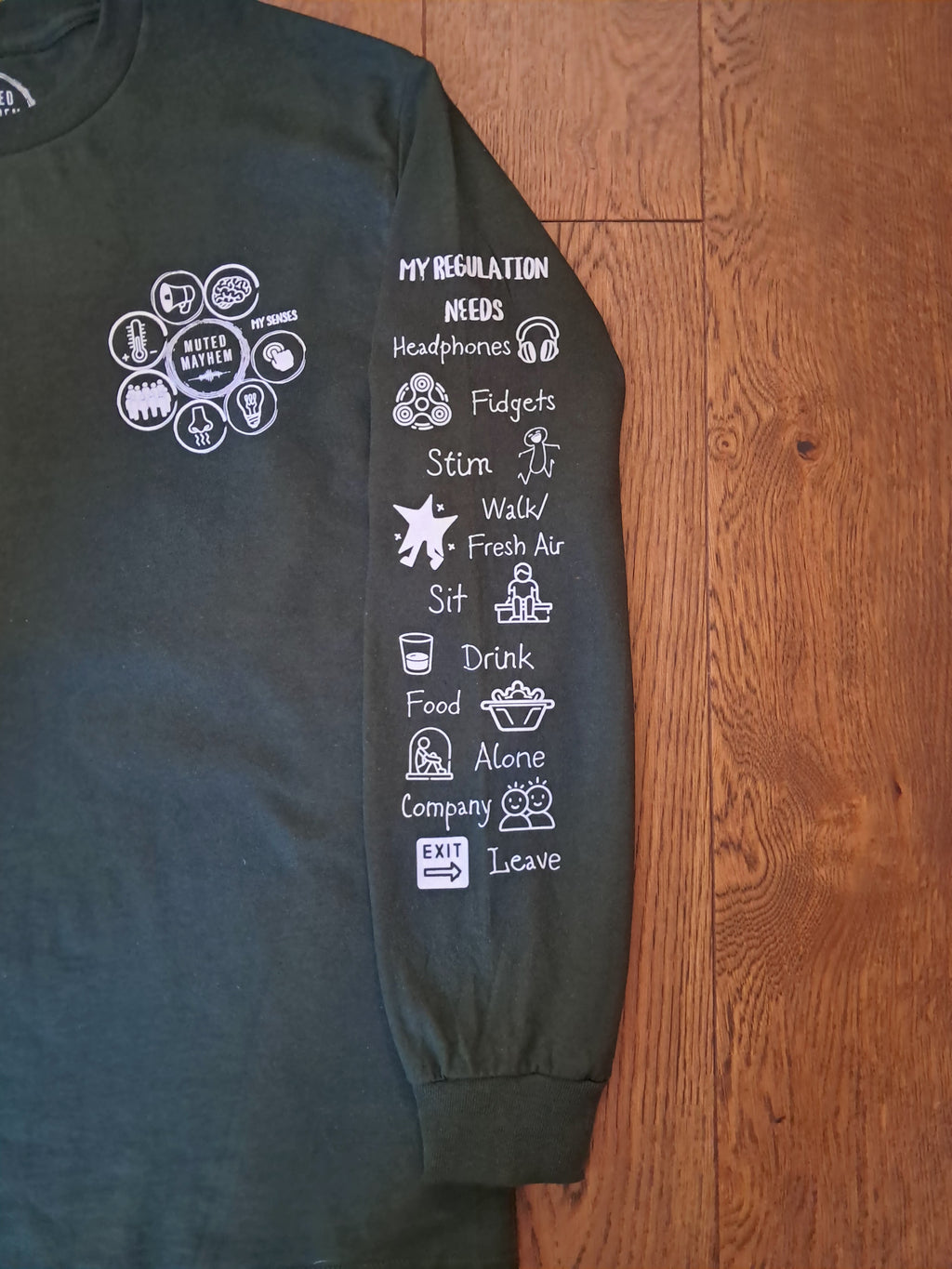 A dark charcoal green long-sleeve t-shirt laid flat on a wooden floor, featuring white printed graphics on both sleeves.