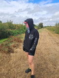 A person standing outdoors on a dry grass path wearing a black hoodie with white graphic prints on the sleeves and chest, black shorts, black sneakers, and a pink cap, with a cloudy sky and greenery in the background.