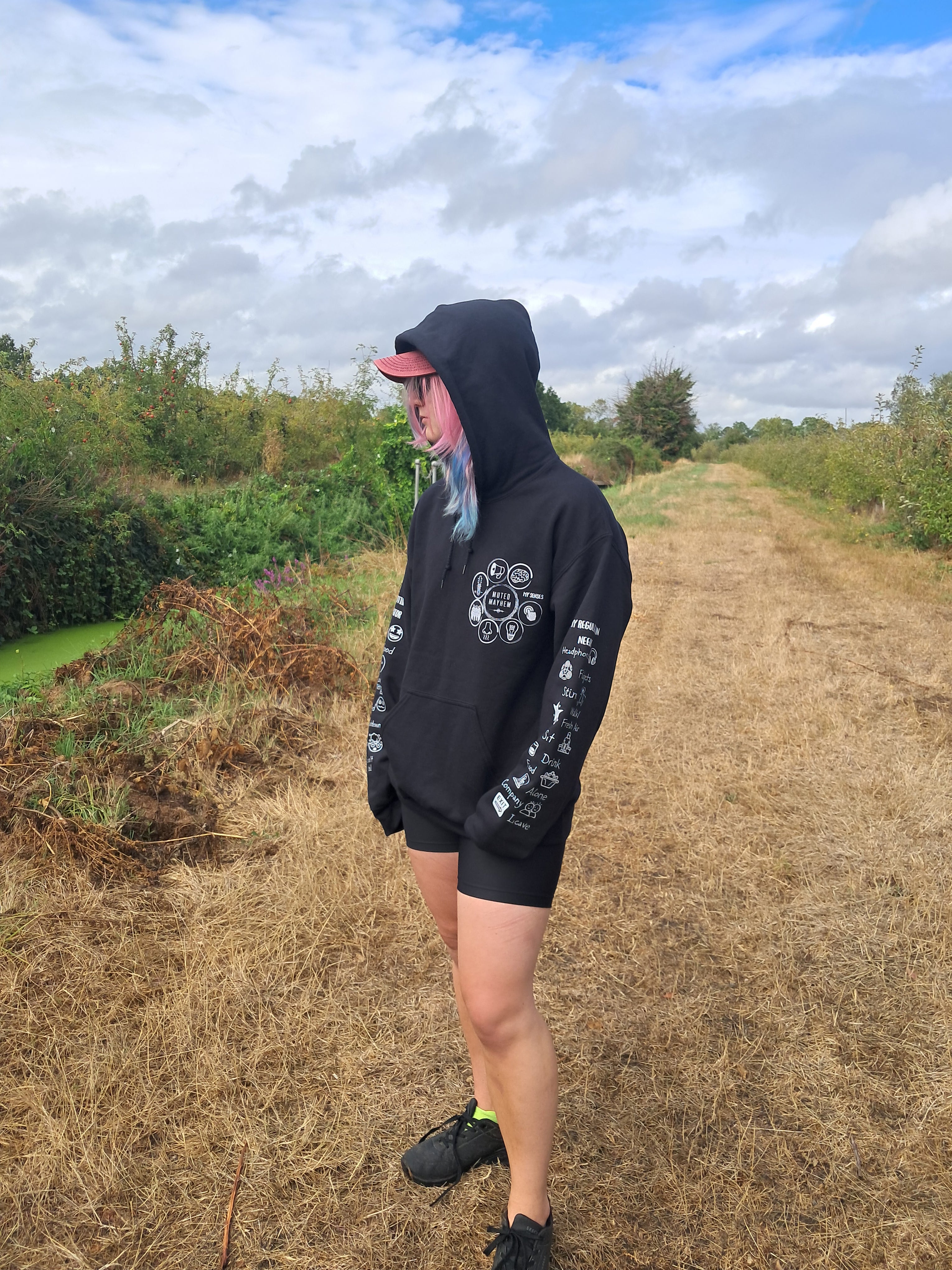 A person standing outdoors on a dry grass path wearing a black hoodie with white graphic prints on the sleeves and chest, black shorts, black sneakers, and a pink cap, with a cloudy sky and greenery in the background.
